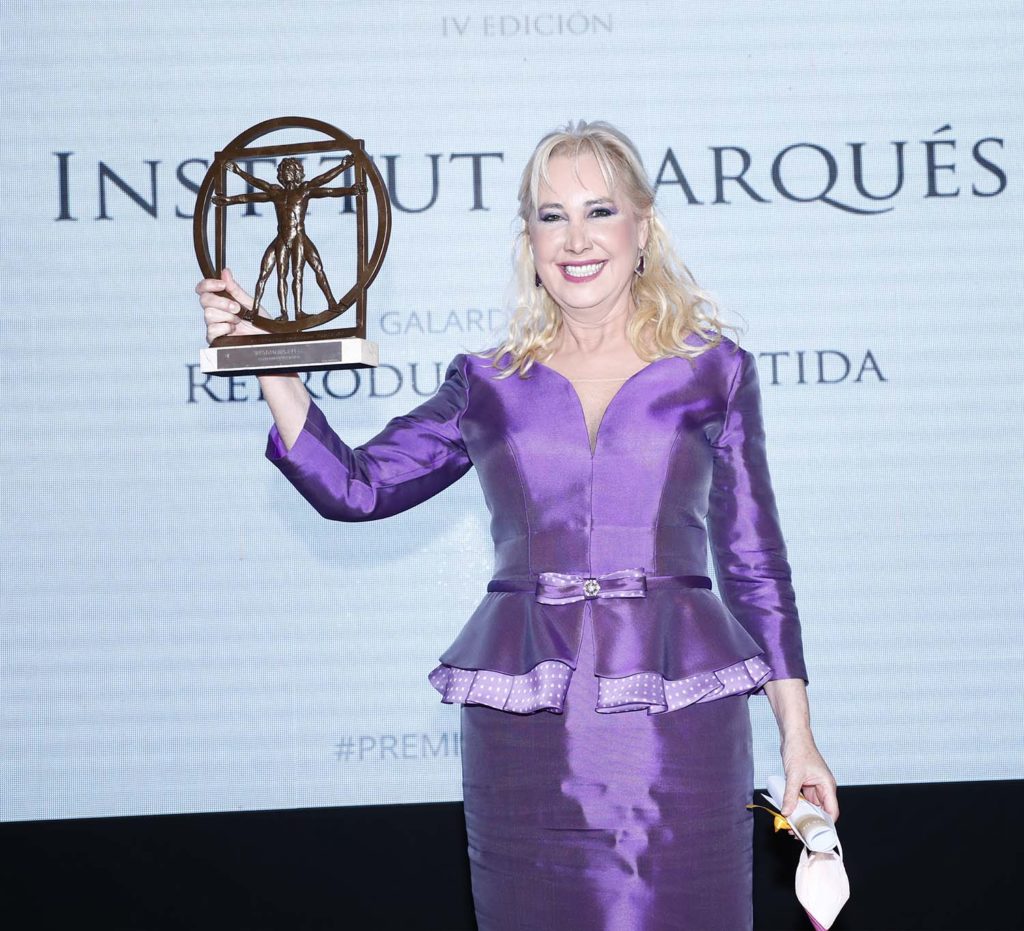 Una mujer con un vestido violeta brillante sostiene un trofeo en forma de estatua del Hombre de Vitruvio y sonríe durante una ceremonia de entrega de premios del Institut Marquès. Está de pie frente a un telón que detalla el evento y con la otra mano sostiene un bolso y unas gafas de sol.